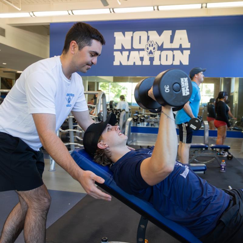 One student lifting weights and another student helping support
